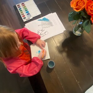 May include: A young child sits at a wooden table, painting with watercolors. They are using a paintbrush and a small blue cup. There is a set of watercolors, a coloring page, and a vase of orange roses on the table.