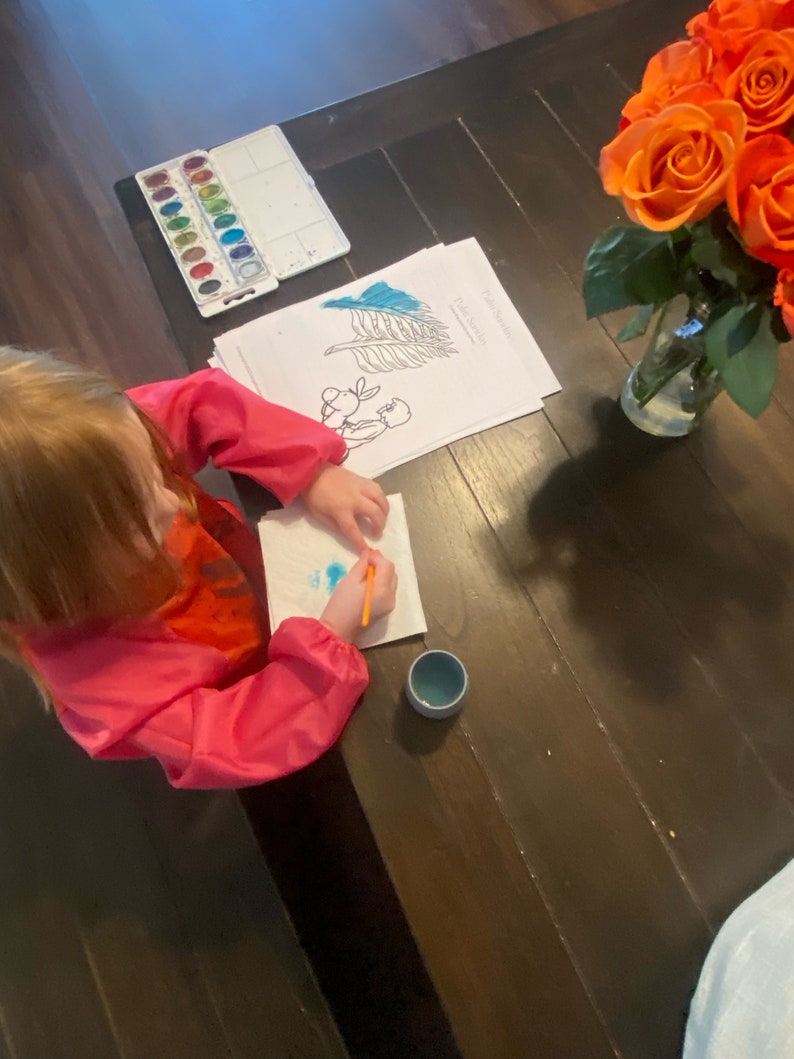 May include: A young child sits at a wooden table, painting with watercolors. They are using a paintbrush and a small blue cup. There is a set of watercolors, a coloring page, and a vase of orange roses on the table.