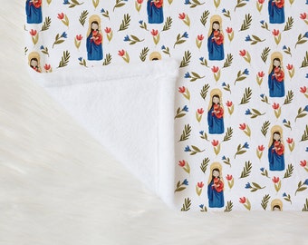 May include: White blanket with a repeating pattern of a religious figure in a blue robe holding a child, surrounded by red and blue flowers and green leaves. The blanket has a soft, white, plush texture.