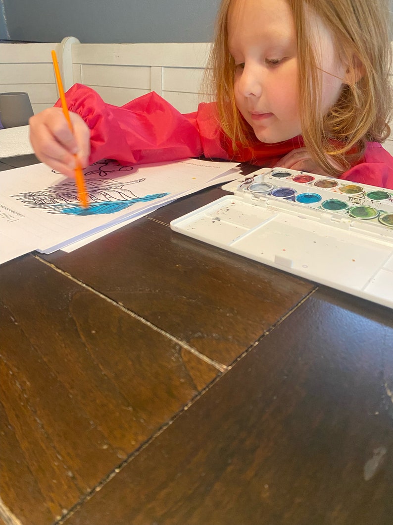 May include: A young person wearing a pink jacket is painting with watercolors. They are using a blue paint to color a picture of a bird. The watercolor set has a white case with a variety of colors.