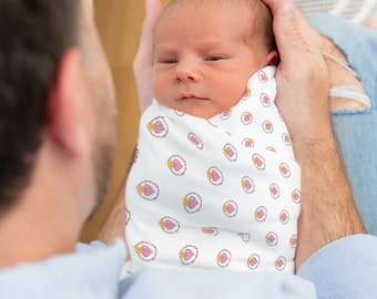 May include: A baby swaddled in a white swaddle blanket with a pink heart and gold crown pattern. The baby is being held by a person.