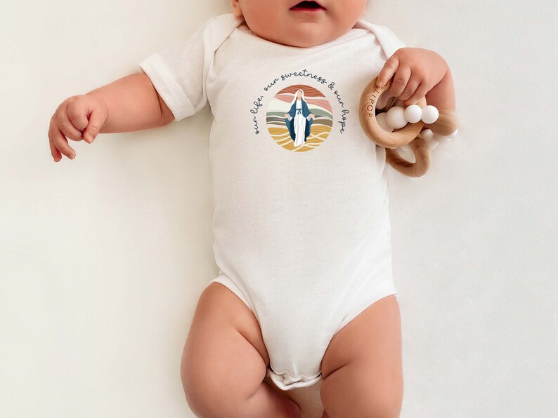 Pu&ograve; includere: Body bianco per neonato con un'illustrazione colorata della Vergine Maria e il testo "our life our sweetness & our hope".