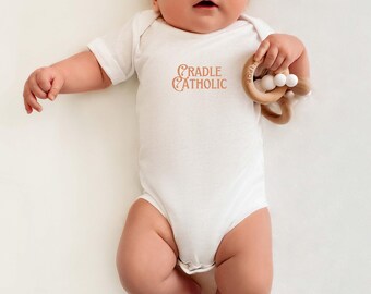 May include: A white baby onesie with the text "CRADLE CATHOLIC" printed in orange on the front. A baby is wearing the onesie and holding a wooden teether.