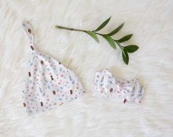 May include: A white baby hat and headband with a floral pattern featuring small illustrations of girls. The pattern includes pink flowers, blue leaves, and white accents.