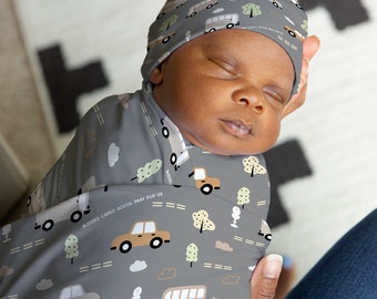 May include: A gray swaddle blanket with a pattern of cars, buses, trees, and clouds. The text "Blessed Carlo Acutis Pray for Us" and "The Eucharist is my highway to heaven" is printed on the blanket. A baby is wearing a matching gray hat with the same pattern.