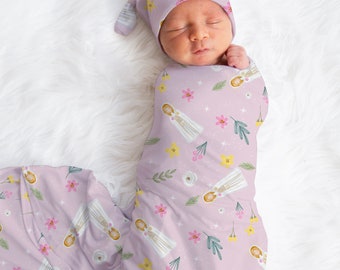 May include: A newborn baby swaddled in a lavender swaddle with a floral pattern featuring white figures and yellow, pink, and green flowers. The baby is wearing a matching lavender hat with a floral pattern.