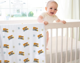 May include: A white baby blanket with a repeating pattern of yellow and gray toy cameras and blue and tan stars. The blanket is draped over a white crib. A baby in a cream-colored onesie is smiling in the crib.
