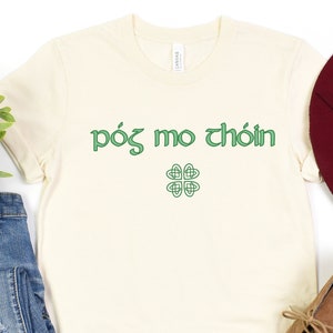 May include: A white t-shirt with the text "póg mo thóin" in green lettering and a green shamrock design. The shirt is laid flat on a white background with a pair of blue jeans, a brown hat, and brown boots.