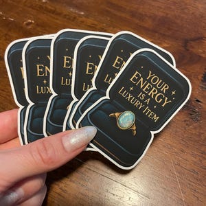 May include: A stack of stickers featuring a ring in a jewelry box. The ring has a light blue opal stone and gold band. The text on the sticker reads "YOUR ENERGY IS A LUXURY ITEM."