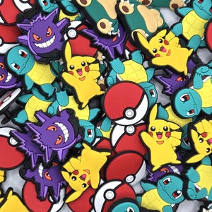 May include: A collection of colorful shoe charms featuring popular cartoon characters. The charms include yellow Pikachu, purple Gengar, blue Squirtle, and red and white Pokeball designs. These rubber charms are a fun accessory.
