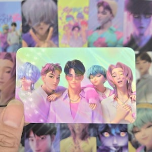 May include: A holographic trading card featuring a group of animated characters with colorful hair and pastel clothing. The card has a gradient background and is held by a hand. Other cards with similar designs are in the background.