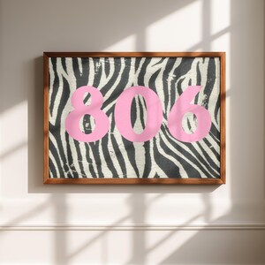 May include: A framed artwork featuring the number "806" in pink against a zebra print background. The artwork is encased in a brown wooden frame, creating a striking contrast. The zebra pattern is black and white.