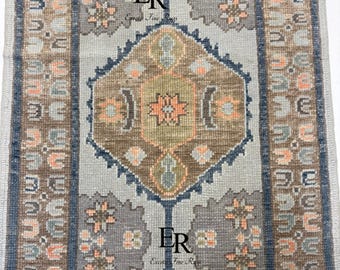 May include: A rectangular rug with a central geometric pattern in shades of grey, brown, and peach. The design features repeating diamond and floral motifs, framed by a border of similar colours. The rug has a vintage, handcrafted appearance.