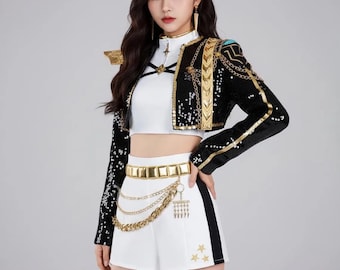 May include: A black cropped jacket with gold sequin trim, gold chains, and shoulder epaulets, worn over a white mock-neck crop top. The outfit includes white high-waisted shorts with a gold metallic belt, hanging gold chains, and three small gold star appliques.