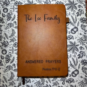 May include: A brown leather journal with the text "The Lee Family" at the top. Below, the journal reads "ANSWERED PRAYERS" and "Psalms 77:11-12." A ribbon bookmark is attached. The journal is on a floral patterned background.