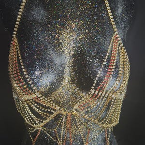 Pode incluir: Uma corrente de corpo dourada com joias e detalhes de cristal vermelho e transparente. O top estilo halter e as mechas em cascata são exibidos em um manequim preto. Joalheria corporal.