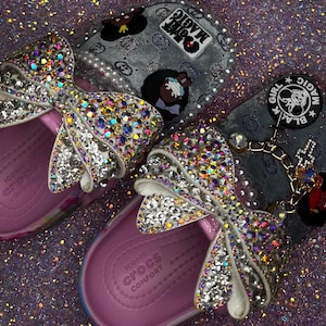 Op de afbeelding: Een paar lichtroze Crocs-slippers met grote strikken bedekt met iriserende strass-steentjes. De bovenste banden zijn versierd met witte parels, bedels en ronde zwart-witte emblemen met de tekst Black Girl Magic in witte letters.