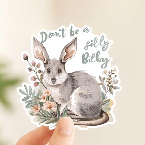 Peut inclure: Un autocollant découpé représentant un bilby gris aux grandes oreilles, entouré de fleurs aux tons doux et de feuilles vertes. Le texte "Don't be a silly Bilby" est imprimé en écriture cursive vert sauge au-dessus de l'animal sur fond blanc.