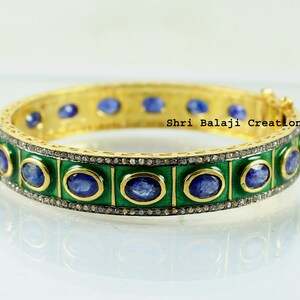 Può includere: Un braccialetto rigido dorato con una fascia in smalto verde e pietre preziose blu ovali. Il braccialetto è decorato con piccole pietre scintillanti. Il testo "Shri Balaji Creations" è visibile.