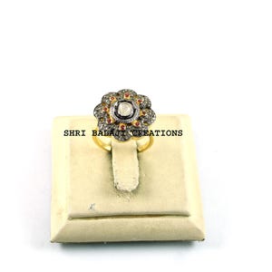 Puede incluir: Un anillo dorado con un diseño en forma de flor. El anillo presenta una piedra central transparente, rodeada de piedras oscuras más pequeñas y detalles rojos. Se exhibe sobre un soporte crema sobre un fondo blanco. El texto "SHRI BALAJI CREATIONS" es visible.