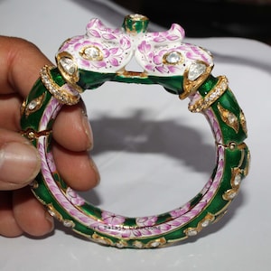 Peut inclure: Un bracelet jonc articulé orné de deux têtes d'éléphant se rejoignant au sommet, fini en émail vert foncé et blanc avec des motifs floraux roses et des pierres claires incrustées. Le bracelet est décoré d'accents dorés et de petits cristaux.