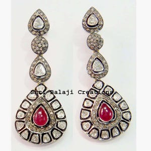 May include: A pair of drop earrings featuring a vertical arrangement of three teardrop and circular shapes set with small clear stones. The base of each earring is a large teardrop shape with a central red stone. Text reads Shri Balaji Creations.