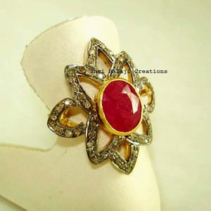 Puede incluir: Un anillo con una piedra preciosa roja redonda central engastada en oro, rodeada por un marco en forma de flor incrustado con pequeñas piedras grises. El texto Shri Balaji Creations es visible en la superficie metálica del anillo.