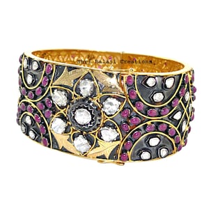 May include: A gold-toned hinged bangle bracelet with a floral design. The bracelet features a central flower with clear stones, surrounded by smaller red stones and gold leaf accents. The text "Shri Balaji Creations" is visible.