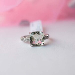 Peut inclure: Une bague argentée ornée d'une grande pierre précieuse vert pâle taillée en coussin au centre. L'anneau est incrusté de multiples petites pierres claires et scintillantes qui s'étendent sur les côtés, créant un aspect brillant et réfléchissant.