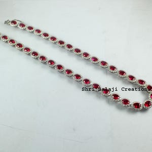 May include: A silver chain necklace with oval-shaped red gemstones surrounded by smaller clear gemstones. The necklace is on a white background.