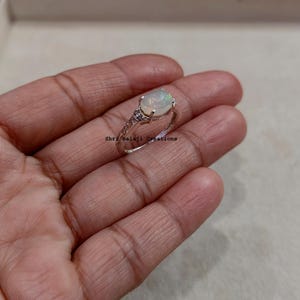 Peut inclure: Une bague en argent ornée d'une pierre d'opale blanche iridescente de forme ovale, fixée dans une monture à quatre griffes. L'anneau présente des motifs métalliques complexes sur les côtés. Le texte Shri Balaji Creations est visible.