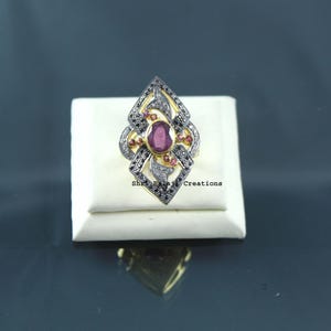 May include: A gold ring featuring a central oval-cut red gemstone surrounded by a geometric frame encrusted with small black stones and smaller red accents. The text Shri Balaji Creations is printed in black on the white display base.