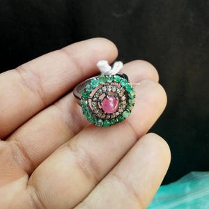 Könnte beinhalten: Ein Ring mit einem zentralen, im Cabochon-Schliff geschliffenen rosa Stein, umgeben von einem kreisförmigen Rahmen aus kleinen klaren Steinen und einem äußeren Rand aus grünen Steinen in einer dunklen Metallfassung. Ein kleines weißes Etikett ist befestigt.