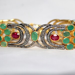 Puede incluir: Una pulsera dorada con varias filas de piedras verdes redondas, dos piedras rojas ovaladas y dos secciones metálicas curvas incrustadas con pequeñas piedras claras. El texto Shri Balaji Creations es visible en la parte derecha.