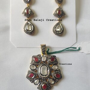 May include: A set of gold-toned jewelry, including a pendant and earrings. The pendant is a floral design with clear and red stones. The earrings have a similar design. The text "Shri Balaji Creations" is visible.