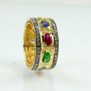 May include: A gold band ring with a row of colorful gemstones. The ring is set with a red ruby, a green emerald, a blue sapphire, and a smaller blue gemstone. The ring is also set with small diamonds.