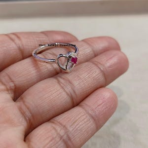 Peut inclure: Une bague argentée dotée d'un sertissage en forme de cœur avec une petite pierre précieuse rose vif et ronde en son centre. L'anneau est fin et poli, et la surface métallique affiche le texte Shri Balaji Creations en petites lettres noires.