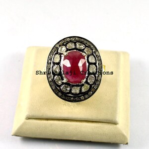 Puede incluir: Un anillo ovalado con una gran gema roja vibrante en el centro, rodeada de piedras más pequeñas y claras engastadas en una banda de metal oscuro. El anillo se exhibe sobre una superficie color crema, mostrando su intrincado diseño.
