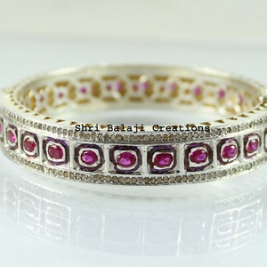 Op de afbeelding: Zilverkleurige armband met een herhalend patroon van vierkant gezette, ovaal geslepen rode edelstenen. De armband is geaccentueerd met kleine, ronde, bruin getinte stenen en heeft de tekst "Shri Balaji Creations".