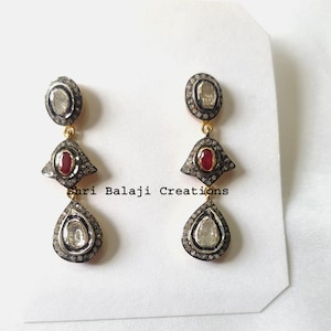 Puede incluir: Pendientes colgantes con tres secciones. La sección superior tiene forma ovalada con una piedra transparente. La sección central tiene forma de diamante con una piedra roja. La sección inferior tiene forma de lágrima con una piedra transparente. El texto "Shri Balaji Creations" es visible.