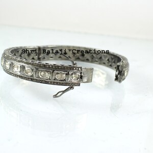 Puede incluir: Un brazalete de plata con una fila de diamantes ovalados engastados en un marco de metal negro. El brazalete tiene un cierre con bisagra y está decorado con pequeños diamantes.