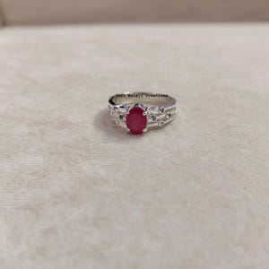 Peut inclure: Une bague en argent avec une pierre rouge centrale de taille ovale. L'anneau est composé de trois fils d'argent parallèles ornés de petites pierres claires rondes. Le texte Shri Balaji Creations est visible au-dessus de la bague.
