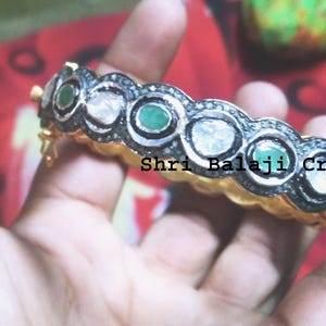 Puede incluir: Una pulsera dorada con engastes alternos de esmeraldas y piedras claras, enmarcados por un borde de metal oscuro. La pulsera se sostiene en una mano, con el texto "Shri Balaji Creations" visible.