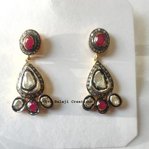 May include: A pair of gold-toned drop earrings. Each earring features a red gemstone at the top, a teardrop-shaped central stone, and three smaller stones at the bottom. The earrings have a black and silver border.