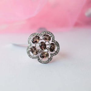 Peut inclure: Une bague argentée avec un sertissage en forme de fleur. Le centre est orné de six pierres précieuses brunes de taille ovale disposées en grappe, entourées d'une bordure de petites pierres claires serties pavé sur le bord des pétales.