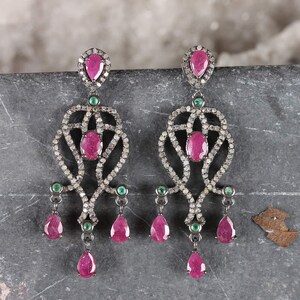 May include: A pair of chandelier earrings featuring dark metal settings encrusted with small pavé stones. Each earring displays multiple pear-shaped and oval-cut pink rubies, accented by small round green emeralds arranged in a symmetrical pattern.