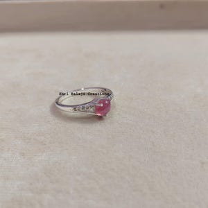 Peut inclure: Une bague en argent avec une pierre rose ronde taillée en cabochon au centre, entourée de petites pierres claires sur l'anneau. Le texte Shri Balaji Creations est imprimé en noir près du sertissage de ce bijou en métal poli et brillant.