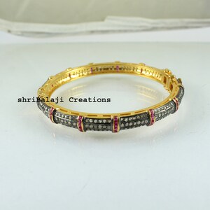 May include: A gold-toned bangle bracelet with a dark gray band encrusted with small, sparkling stones. The bracelet features sections of small red stones and the text "shrilalaji Creations".