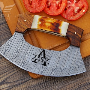 May include: A personalized, stainless steel cleaver with a wooden handle and a bone-like accent. The blade has a Damascus pattern and is engraved with the name "Arther".