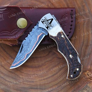 May include: A folding knife with a patterned blade and an antler handle. The blade features a distinctive design with copper accents. The knife is engraved with the name "ARTHER" and is accompanied by a brown leather sheath.